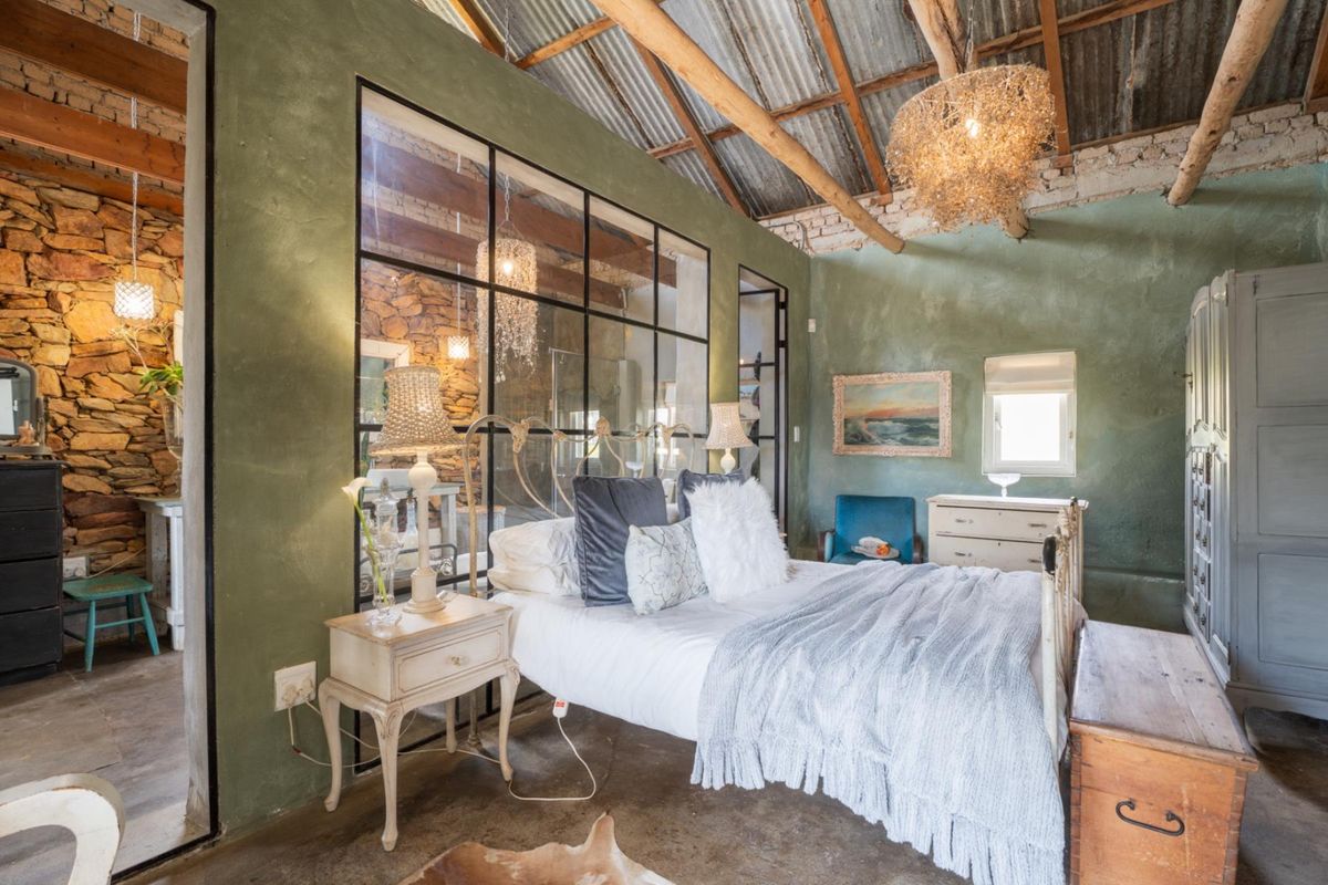 A rustic bedroom featuring exposed timber beams, corrugated iron ceiling, textured green walls, and a large industrial-style steel-framed glass partition.