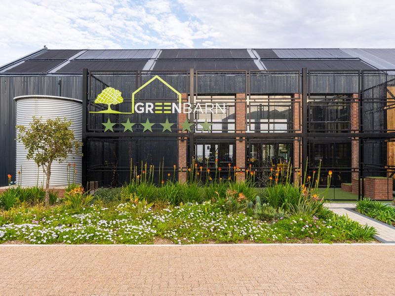 Modern industrial-style building facade featuring corrugated metal cladding, integrated solar panels, a water tank, and decorative metal mesh screening with landscaped garden beds.