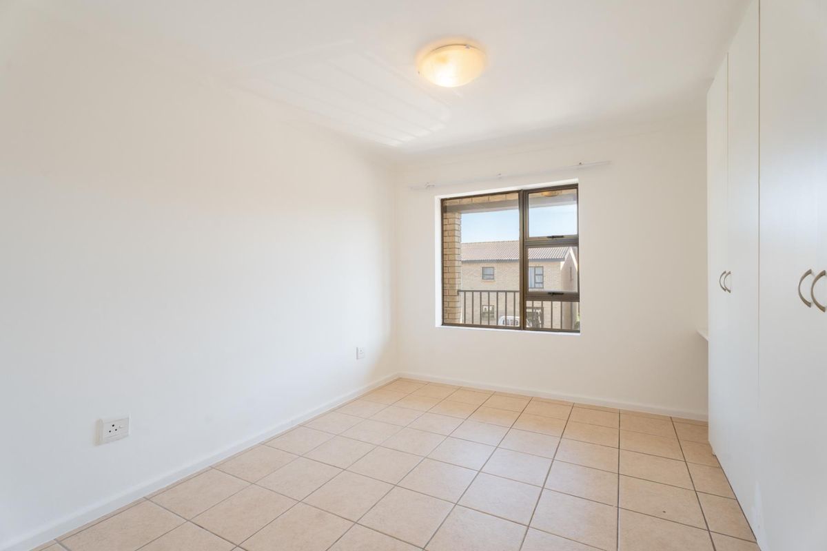 A bright, empty bedroom featuring neutral tiled flooring, built-in cupboards, and an aluminium-framed window providing natural light.