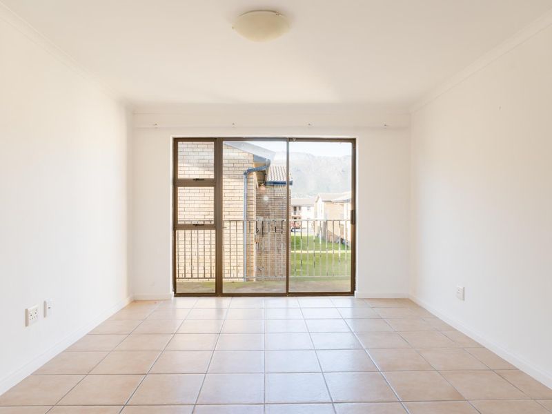 An empty, sunlit living area featuring neutral tiled flooring and a large sliding glass door that opens onto a balcony.