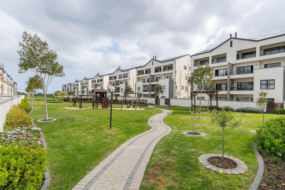A well-maintained communal garden area featuring a paved walkway, landscaped lawns, and views of the apartment complex facades with private balconies.