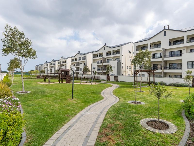 A well-maintained communal garden area featuring a paved walkway, landscaped lawns, and views of the apartment complex facades with private balconies.