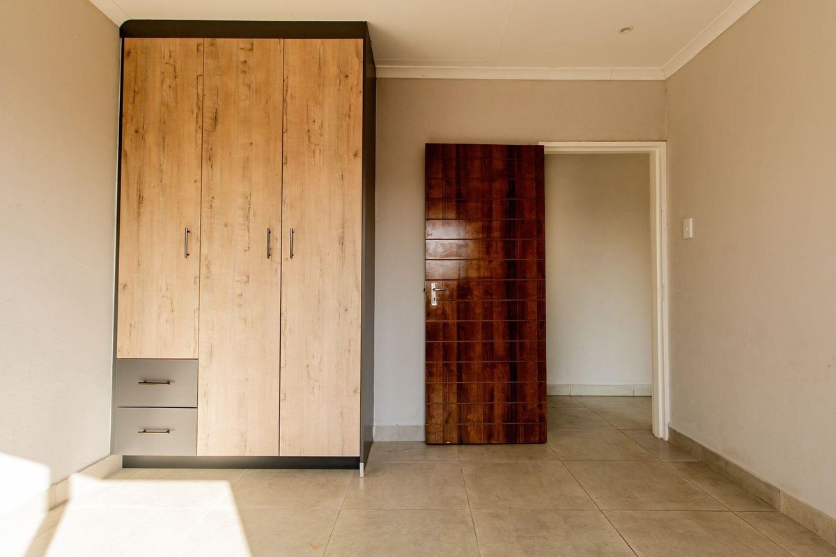 A bright bedroom featuring built-in wooden cabinetry and tiled flooring, leading to an adjacent room through a dark wood door.