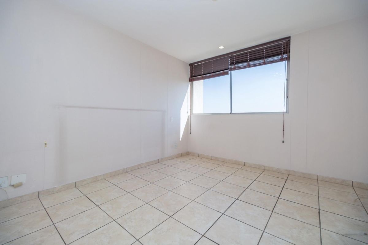A bright, empty bedroom featuring neutral floor tiles, a large window with fitted blinds, and clean white walls.
