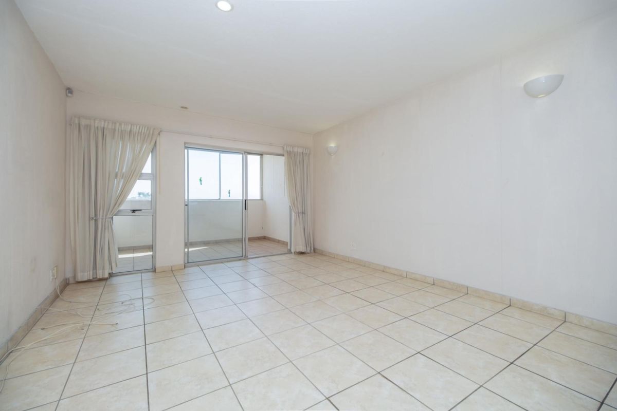 A bright, empty living room featuring neutral ceramic floor tiles, recessed lighting, and sliding glass doors leading to an attached balcony.