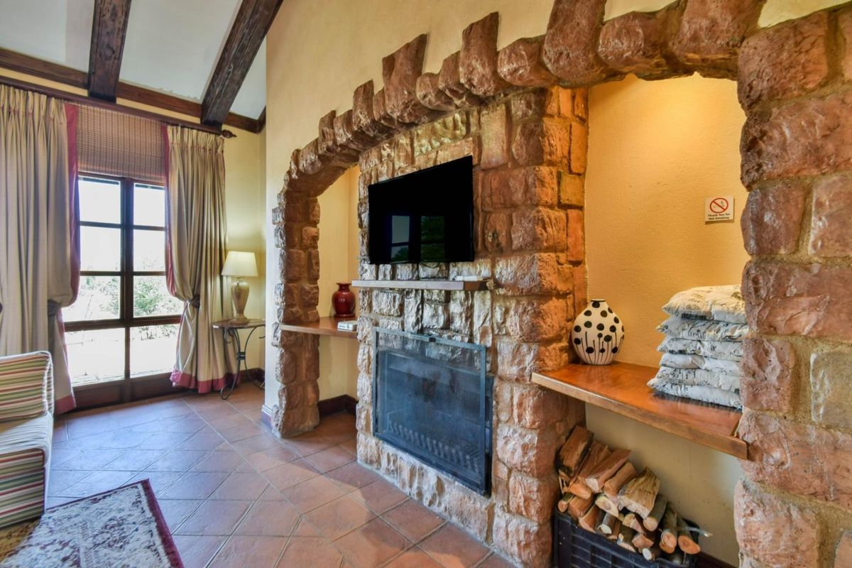A rustic living room featuring a prominent stone fireplace, exposed timber ceiling beams, and terracotta floor tiles, complemented by classic french doors.