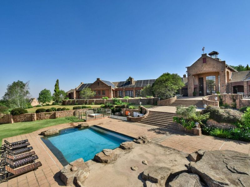 Expansive rear exterior view featuring a stone-clad residence, a swimming pool with stone surround, tiered landscaping, and integrated solar panels on the roof.