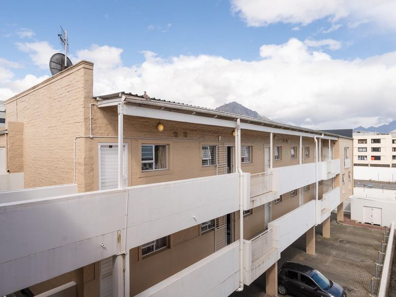 Exterior view of a multi-story apartment building featuring brick facade, balcony walkways with metal railings, and a paved ground-level parking area.