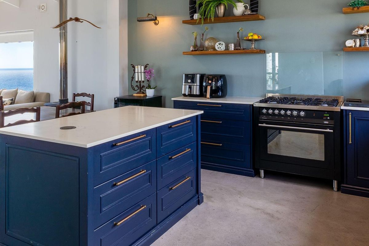 Modern kitchen featuring deep blue cabinetry, stone countertops, a central island, and a professional-style gas range, set against a coastal backdrop.