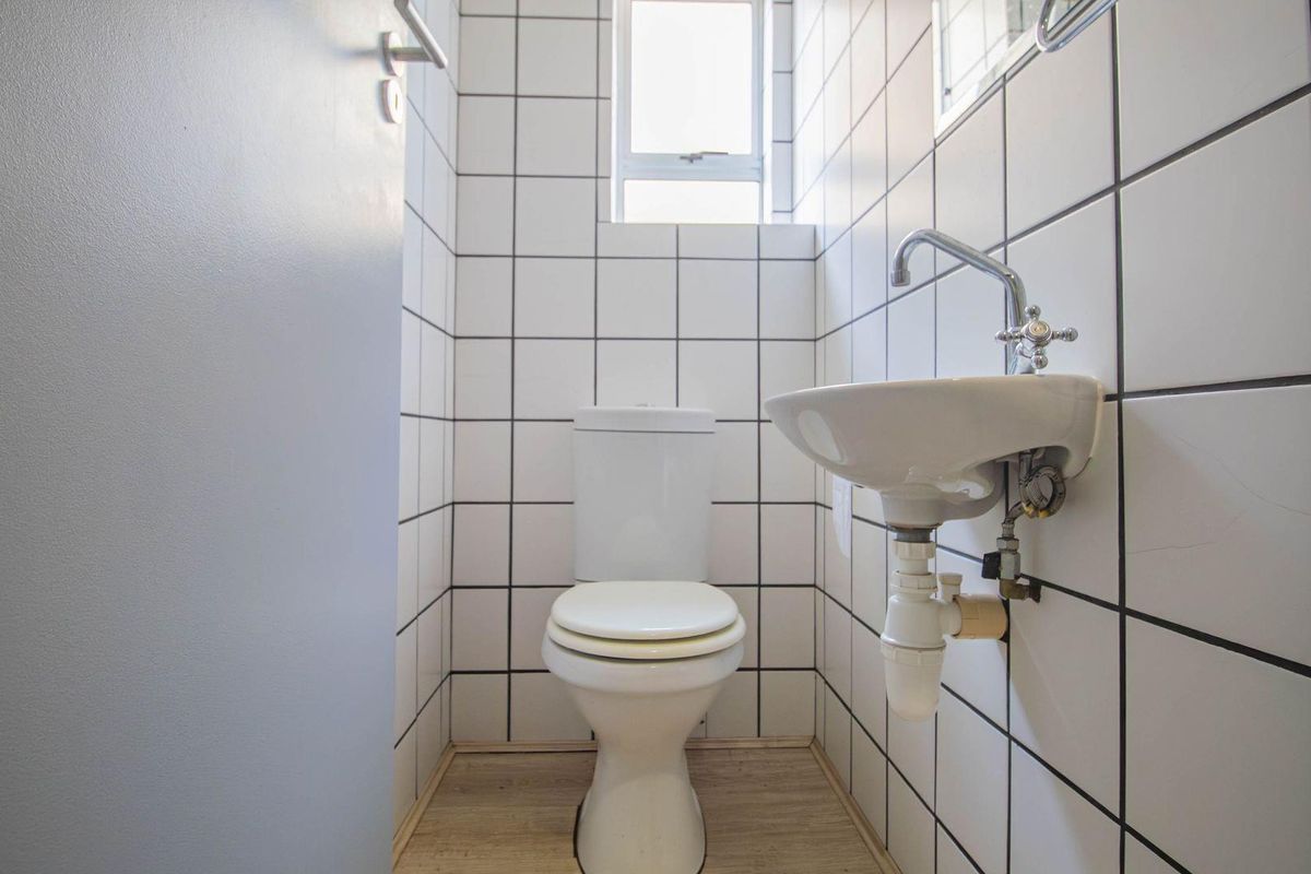 A compact office restroom featuring white wall tiles with dark grout, a wall-mounted basin, and a standard toilet.