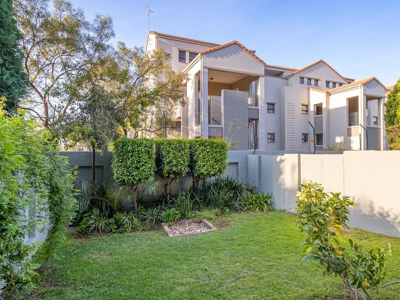 A well-maintained rear garden area featuring a manicured lawn, established landscaping, and boundary walls, set against the backdrop of a multi-story apartment complex.