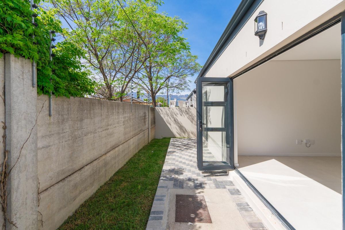 A side garden area featuring paved walkway, concrete boundary wall with electric fencing, and direct access to the interior through modern glass doors.