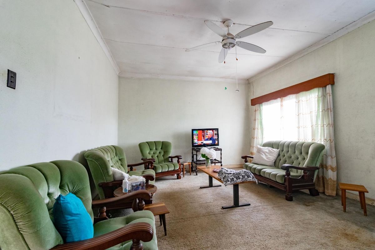 A dated living room featuring a ceiling fan, carpeted flooring, and large windows with a wooden pelmet.