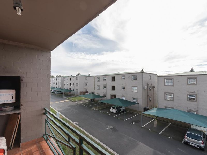A covered balcony featuring a built-in braai, tiled flooring, and metal railings, overlooking the apartment complex parking area.