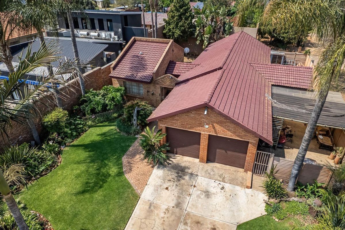 Aerial view of a brick house featuring a double garage, paved driveway, landscaped garden, and a covered patio area.
