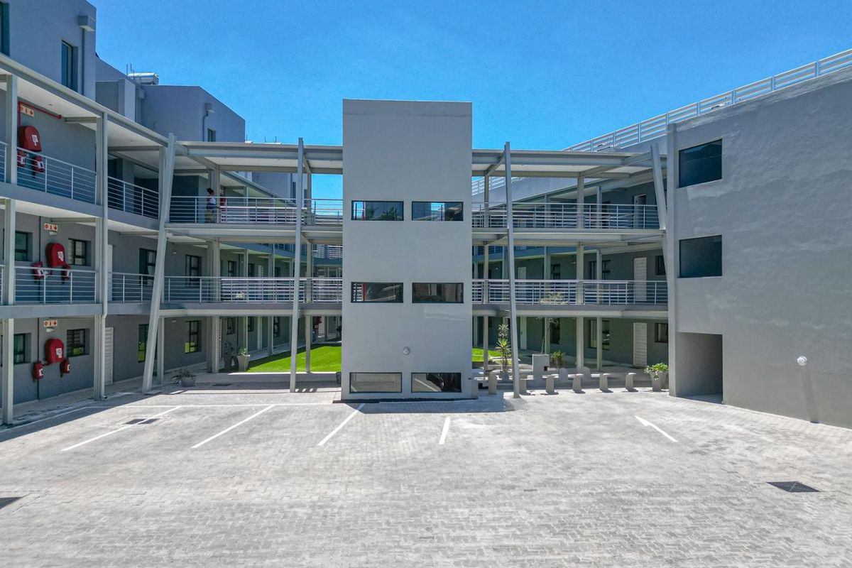 A modern apartment complex exterior featuring a paved parking courtyard, multi-level walkways with metal railings, and a central stairwell tower.