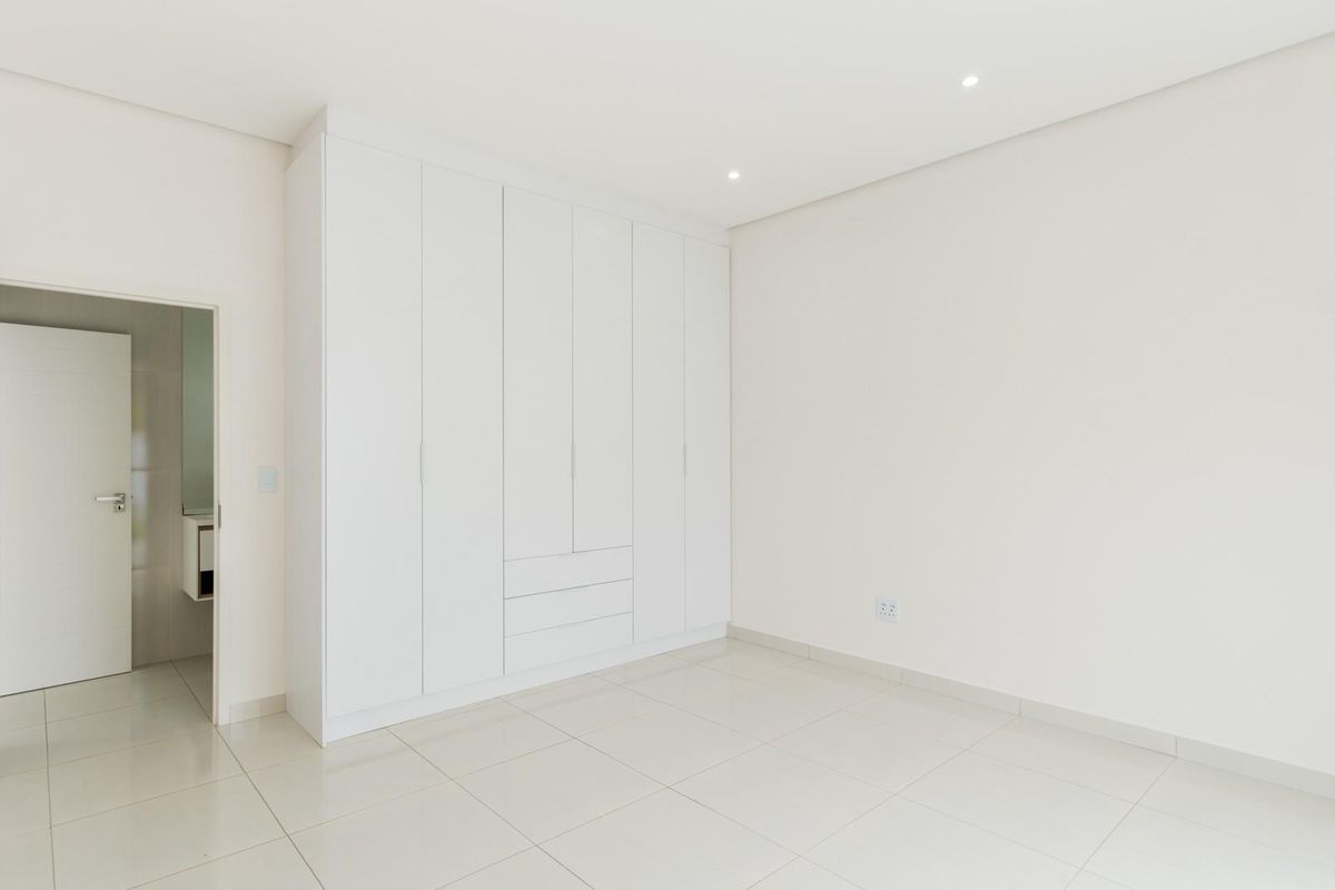 A bright, modern bedroom featuring neutral tiled flooring, recessed ceiling lighting, and extensive built-in white cabinetry with integrated storage.