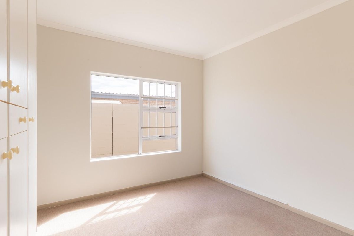 A bright, empty bedroom featuring neutral carpeting, built-in cupboards, and a window with security bars.