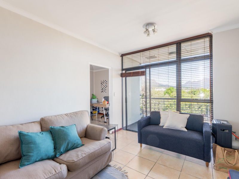 A bright living room featuring tiled flooring, large windows with venetian blinds, and direct access to an outdoor balcony area.