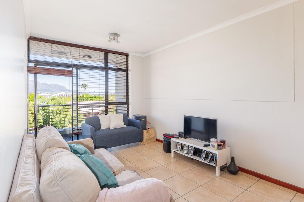 A bright living room featuring tiled flooring, a large sliding glass door with balcony access, and venetian blinds, offering natural light and mountain views.