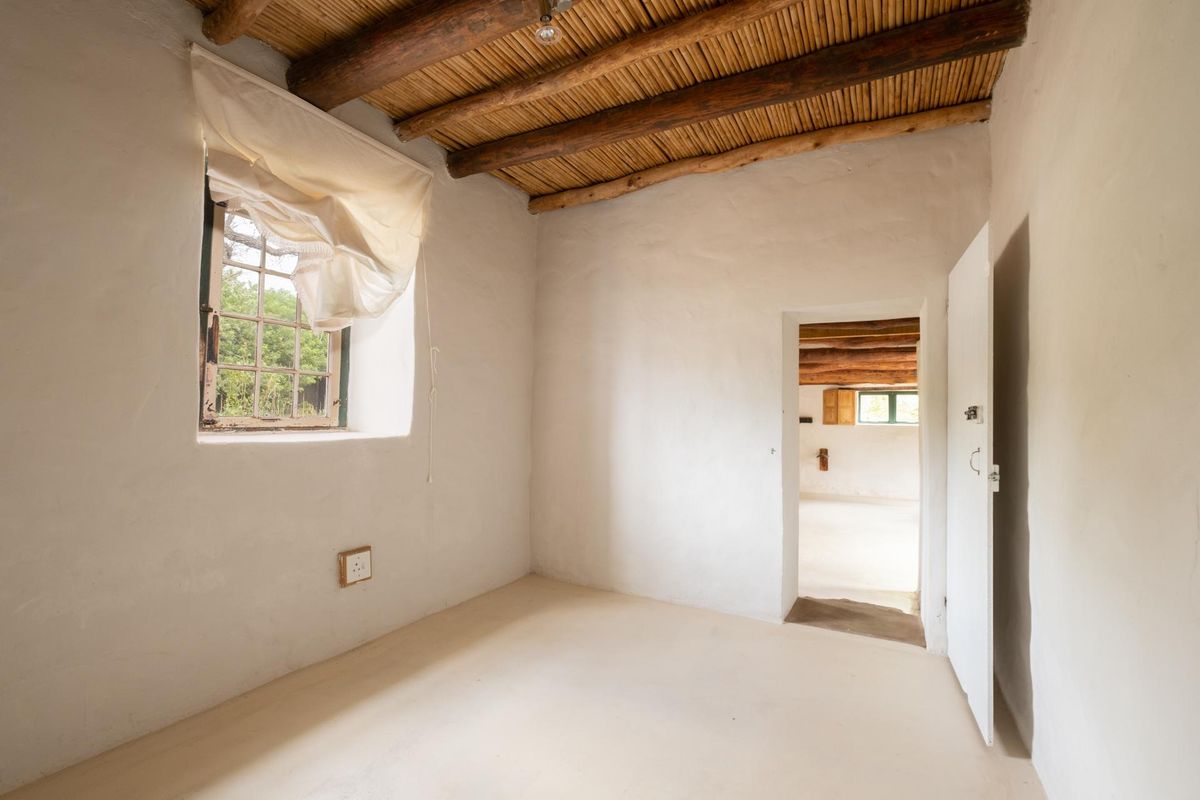 A rustic bedroom featuring white plastered walls, exposed timber ceiling beams with reed infill, and a small window overlooking the exterior.