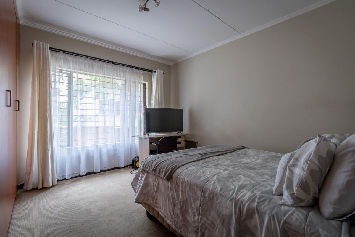A well-lit bedroom featuring neutral carpeting, built-in wooden cupboards, and a large window with security bars, providing a comfortable and functional living space.
