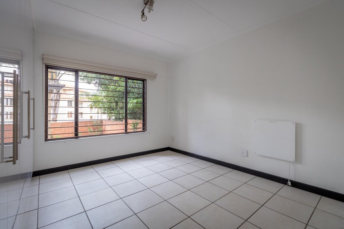 A bright, empty bedroom featuring neutral floor tiles, a large window with security bars, and a wall-mounted electric heater.