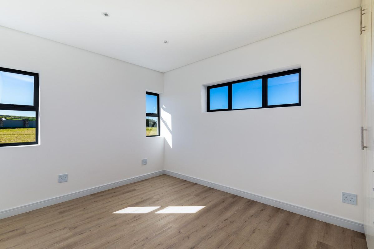 A bright, empty bedroom featuring modern wood-look flooring, clean white walls, and black-framed windows offering natural light and exterior views.