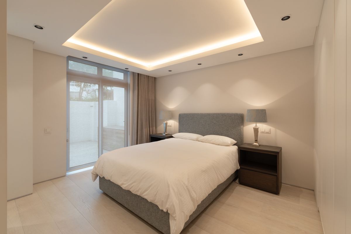 Modern bedroom featuring a recessed tray ceiling with integrated LED lighting, light wood flooring, and a glass door providing access to a private patio.