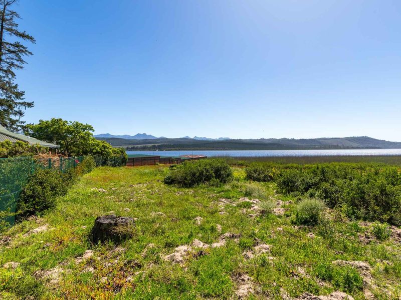 A vacant residential plot featuring natural vegetation, a perimeter fence, and expansive views of the nearby lake and mountain range.