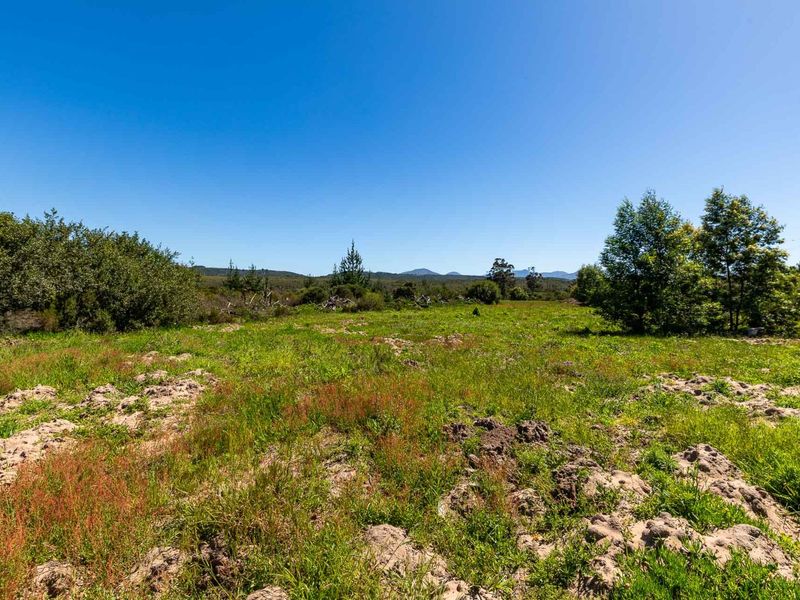 A view of the vacant residential land featuring natural vegetation, uneven terrain, and distant mountain views under a clear blue sky.