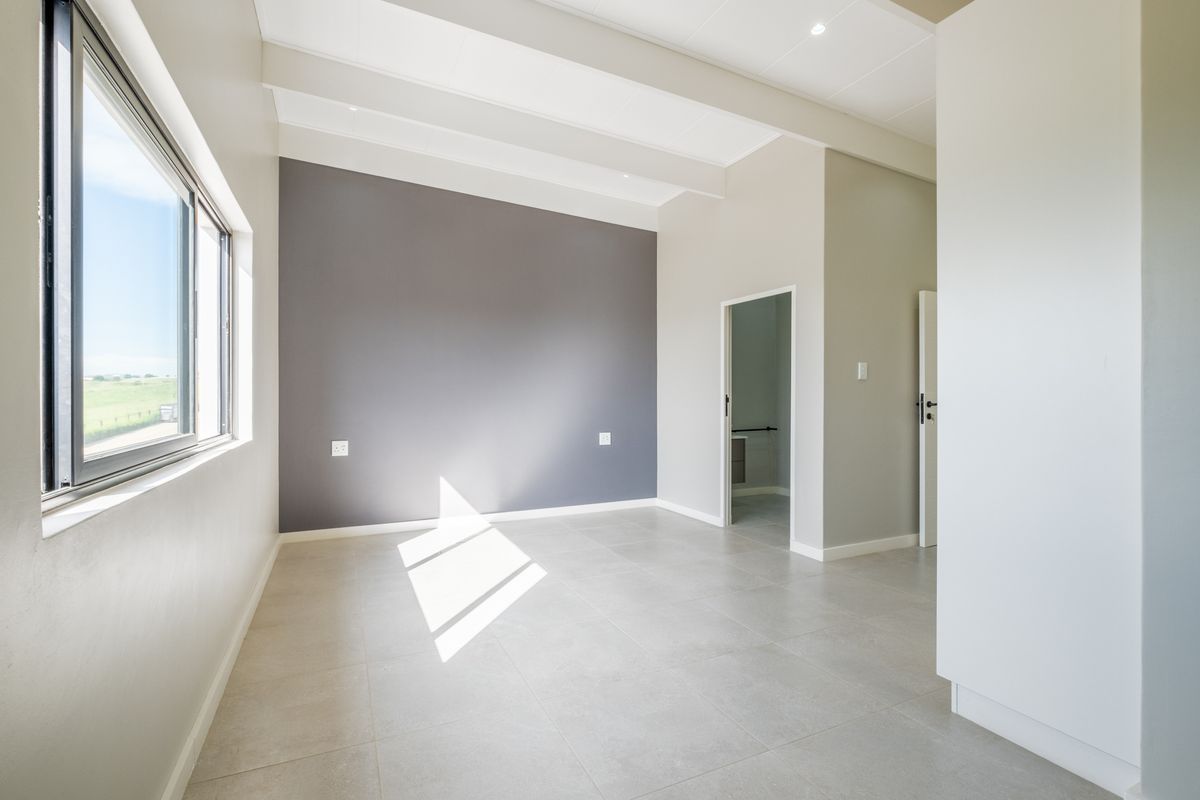 A bright, modern bedroom featuring neutral tiled flooring, a striking grey accent wall, exposed ceiling beams, and large windows providing natural light.
