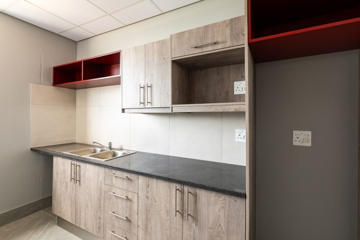 A modern kitchenette featuring wood-grain cabinetry, dark stone countertops, a stainless steel double sink, and integrated shelving with red accents.