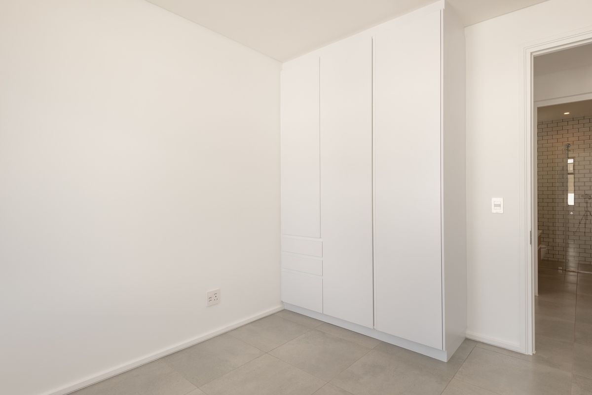 A clean, minimalist bedroom featuring built-in white cabinetry and large-format floor tiles, with an adjacent view into a tiled bathroom.