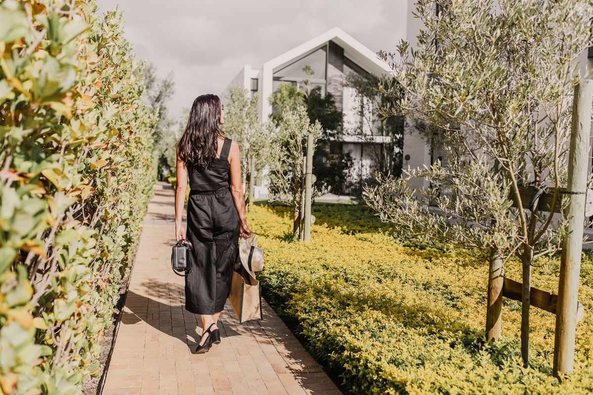 A well-maintained paved walkway lined with manicured hedges and young olive trees leading towards a modern residential building with a gabled roof.