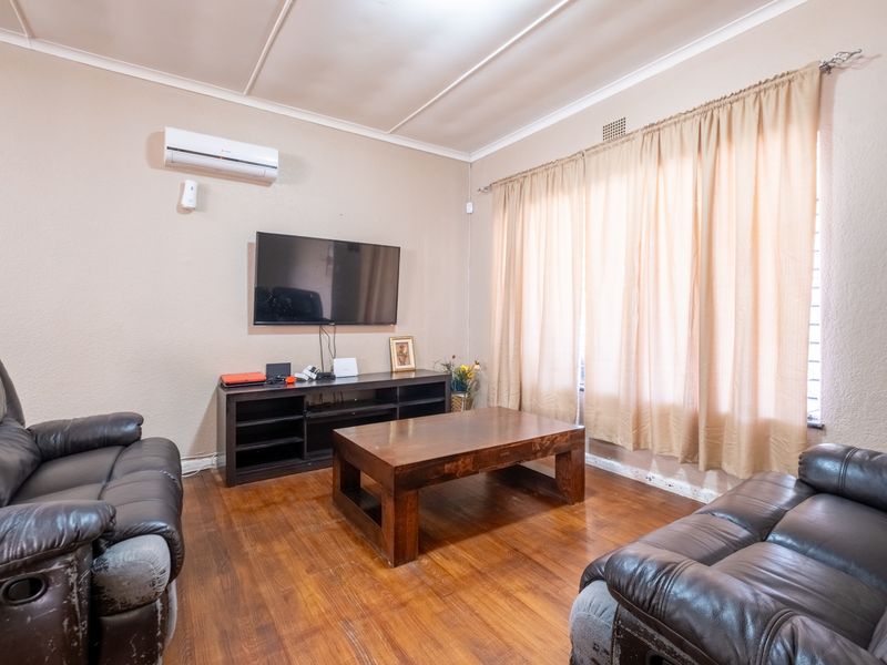 A living room featuring hardwood flooring and a wall-mounted air conditioning unit, with large windows dressed in curtains.