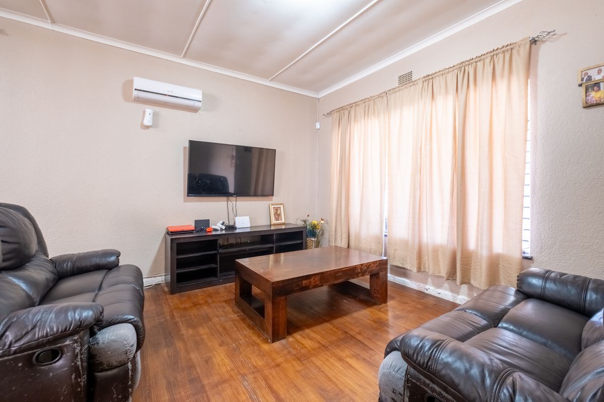 A living room featuring hardwood flooring and a wall-mounted air conditioning unit, with large windows dressed in curtains.