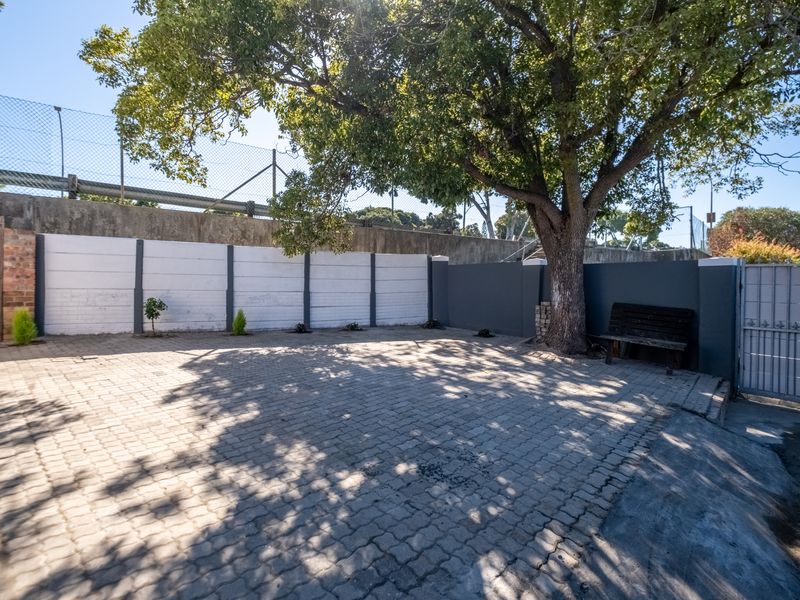 A paved outdoor area featuring a boundary wall, metal gate, and a large mature tree providing shade.
