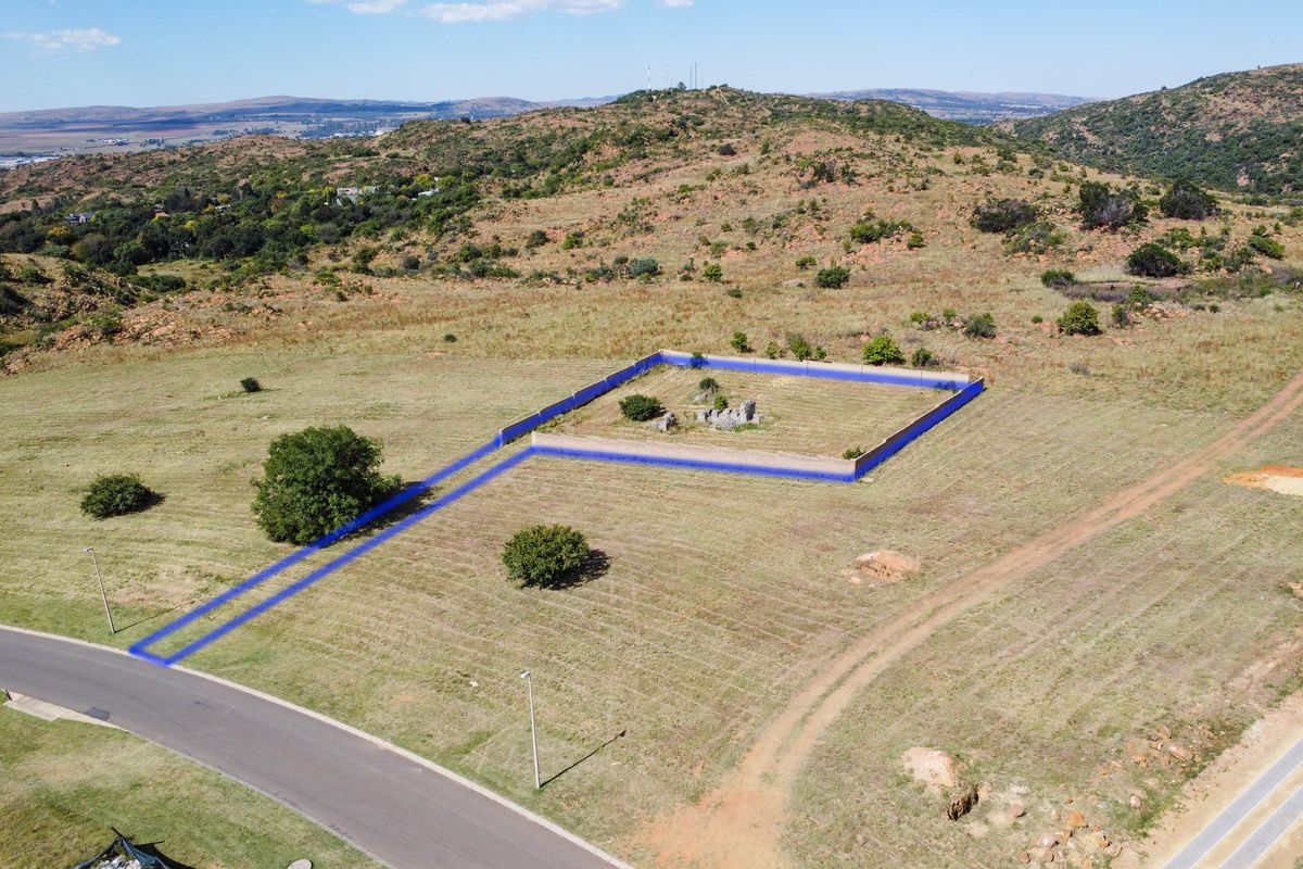 Aerial view of a vacant residential plot featuring a fenced enclosure, natural landscape, and direct access to a paved street.