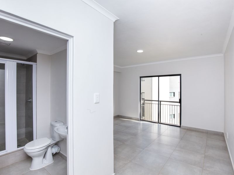A bright, open-plan living area featuring neutral floor tiles, sliding glass doors to a balcony, and an adjacent modern bathroom with a glass shower.