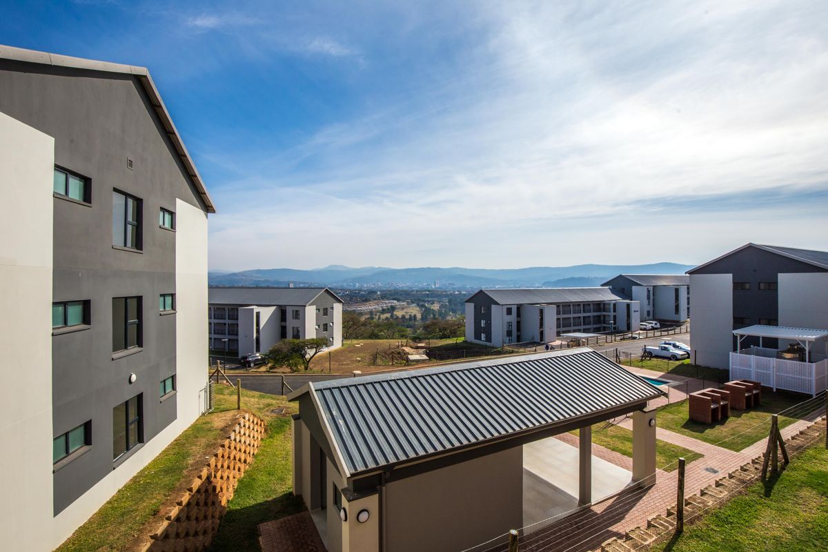Elevated view of the apartment complex grounds, featuring modern multi-story residential buildings, a communal covered patio area, and scenic landscape views.