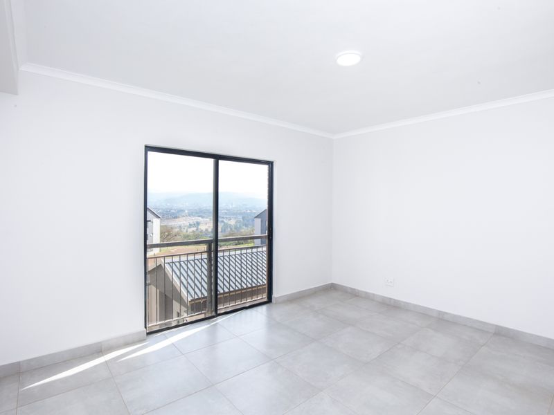 A bright, empty bedroom featuring neutral floor tiles, white walls, and a sliding glass door leading to a balcony with scenic views.