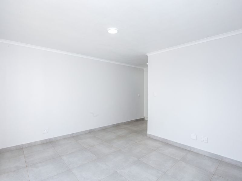 A bright, empty living area featuring neutral tiled flooring, clean white walls, and recessed ceiling lighting, offering a versatile space for customization.