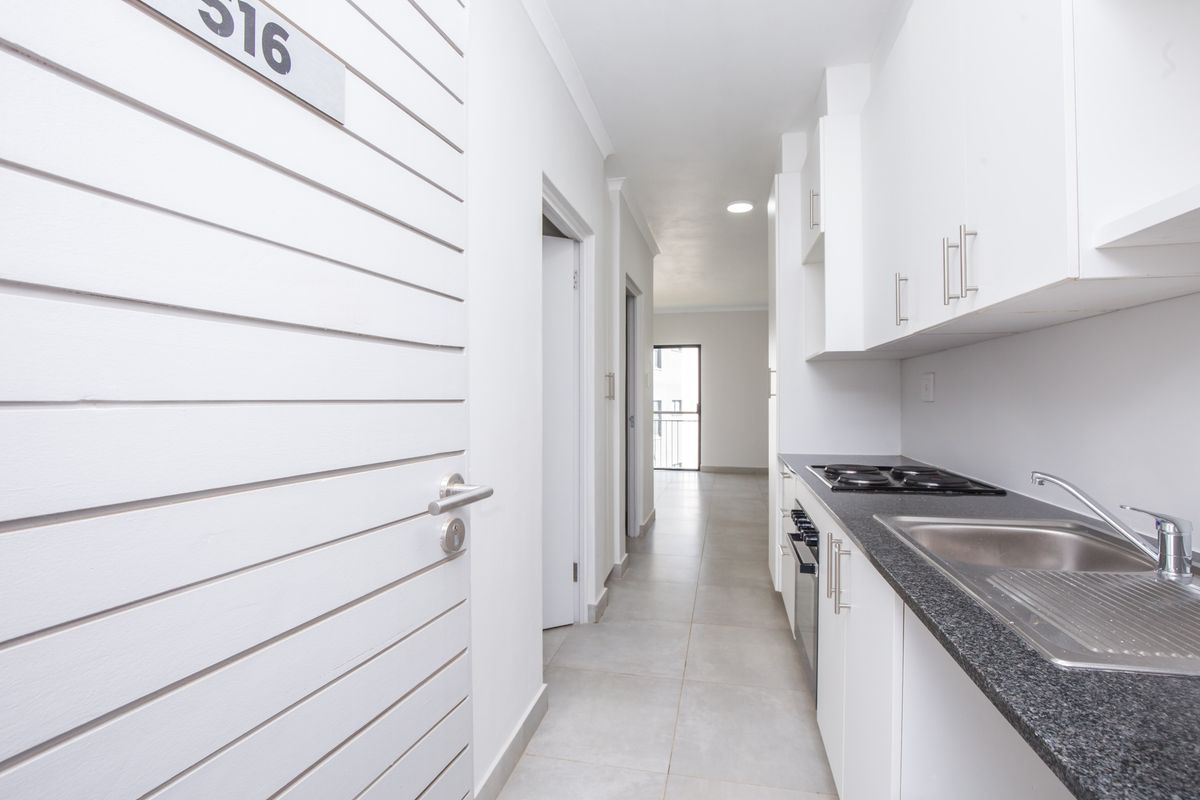 A clean, modern galley-style kitchen featuring white cabinetry, dark granite countertops, a built-in hob, and an integrated sink, leading into a bright hallway.