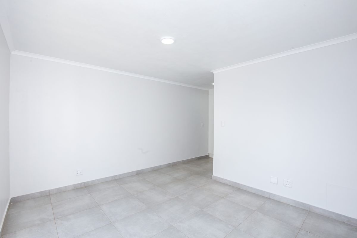 A bright, empty living area featuring neutral tiled flooring, clean white walls, and recessed ceiling lighting, offering a versatile space for customization.