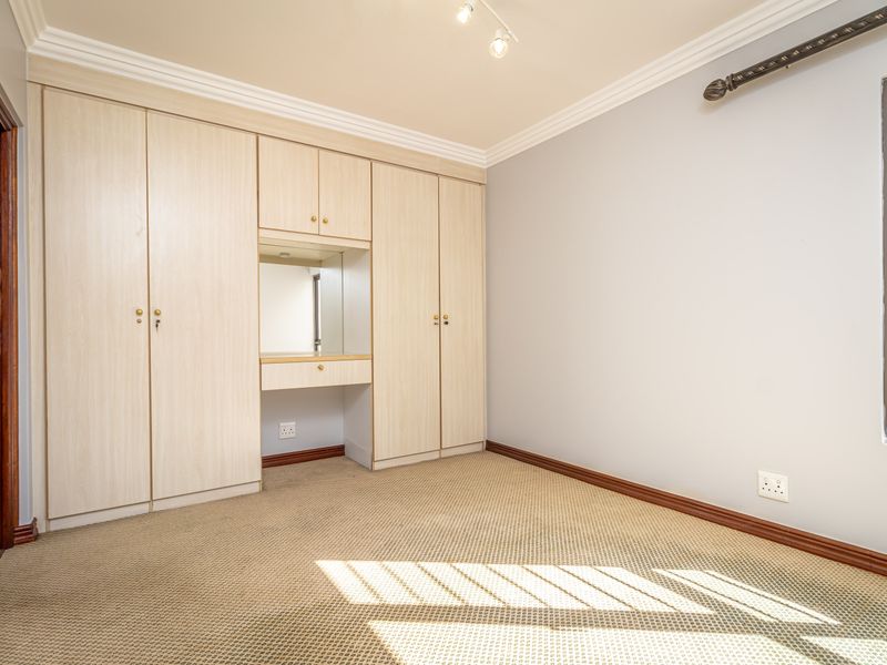 A clean, empty bedroom featuring built-in cupboards with an integrated vanity mirror, carpeted flooring, and security bars on the window.