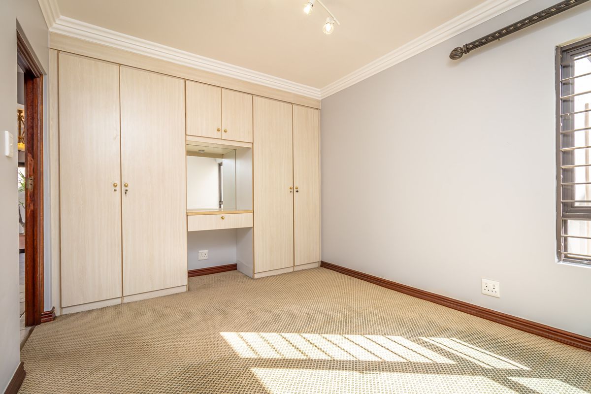 A clean, empty bedroom featuring built-in cupboards with an integrated vanity mirror, carpeted flooring, and security bars on the window.