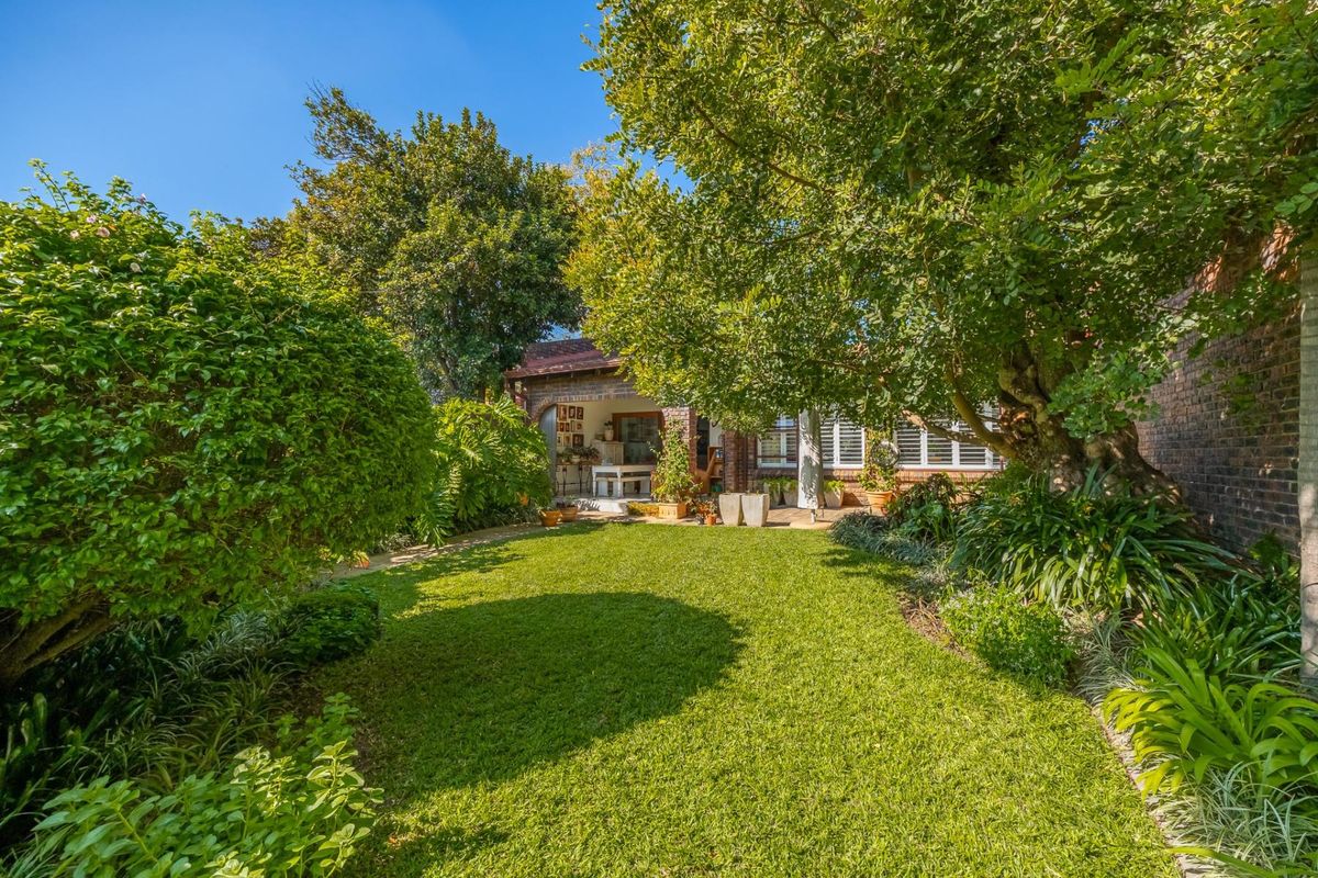 A lush, well-maintained rear garden featuring a manicured lawn, mature trees, and a covered patio area attached to the brick townhouse structure.