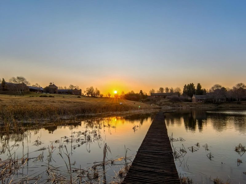 A scenic sunset view over a tranquil lake featuring a wooden jetty, surrounded by expansive landscaped grounds and distant residential properties.