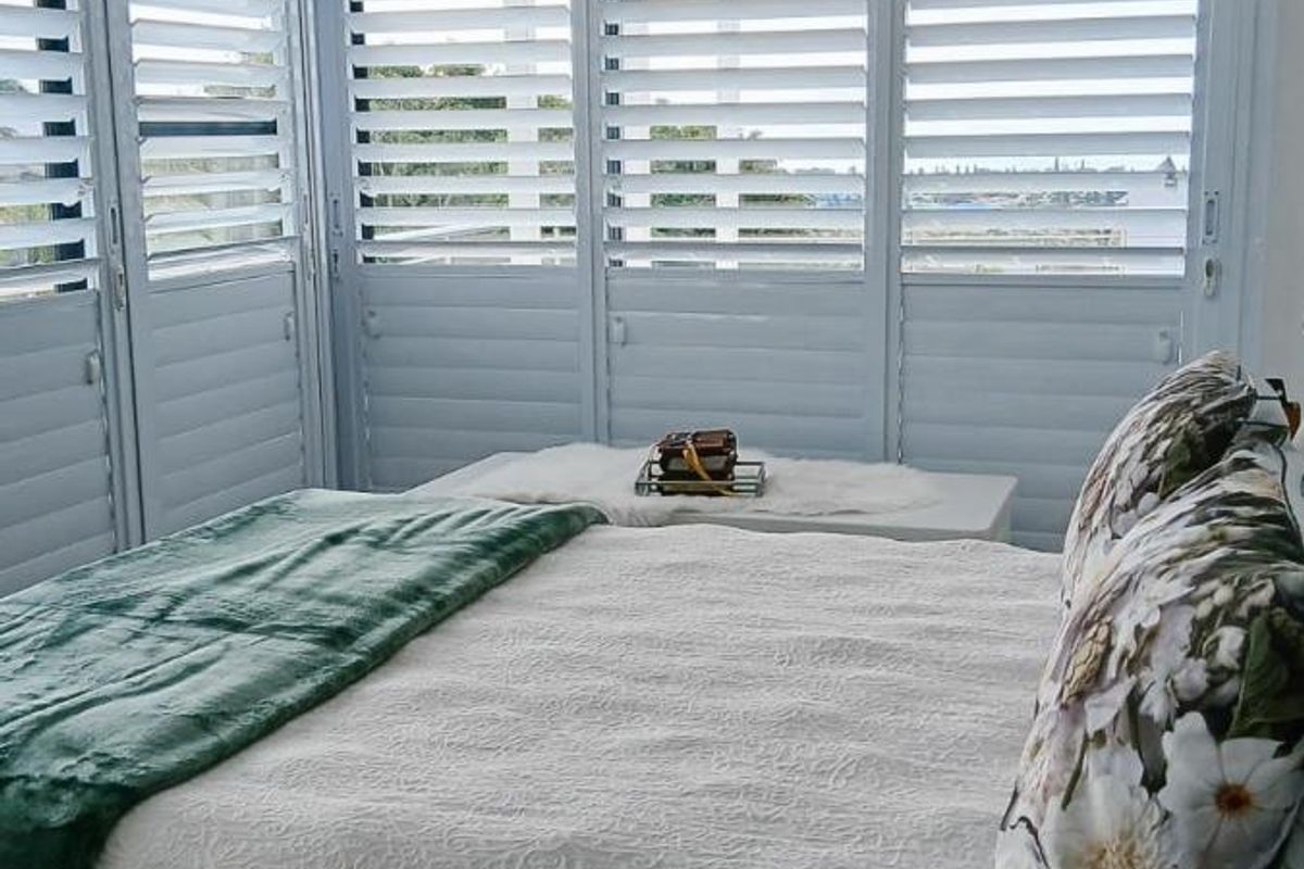A bright bedroom featuring white plantation shutters, a crystal chandelier, and neutral finishes, offering a clean and well-maintained living space.